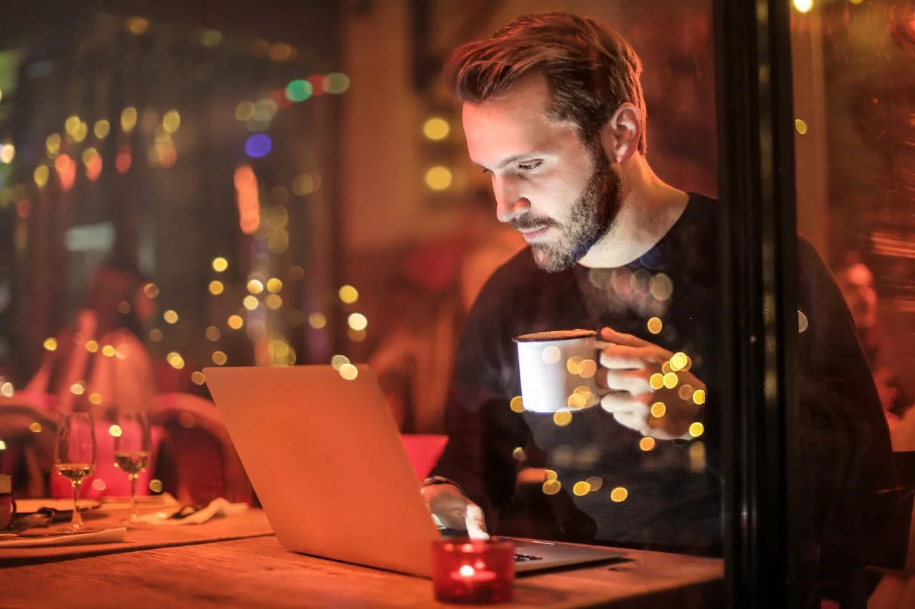 A bearded man working intently on a laptop at night in a warmly lit cafe, holding a mug