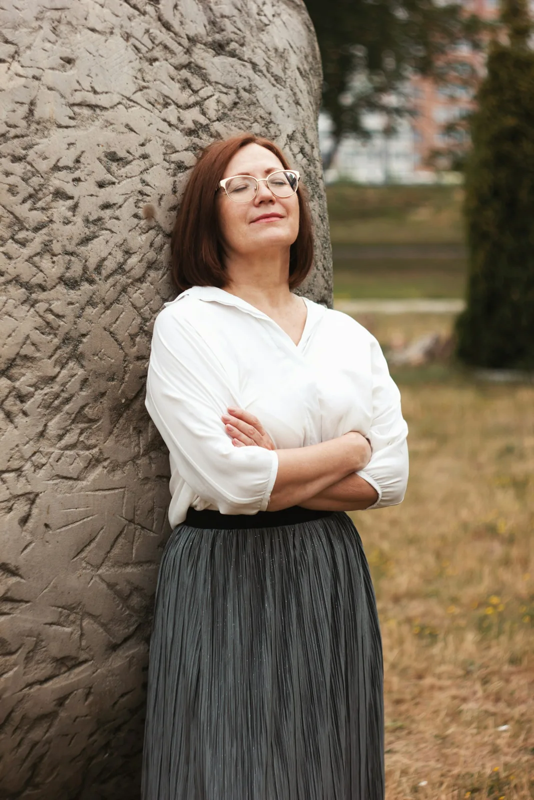 A calm woman in a white blouse leaning against a tree with her arms folded