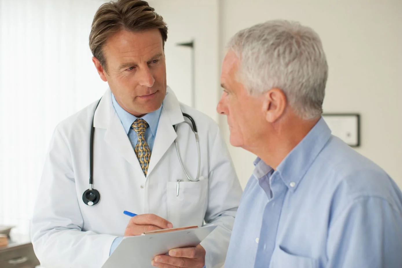 A clinician in a white coat reviewing testosterone treatment options with an older male patient in a bright clinical office