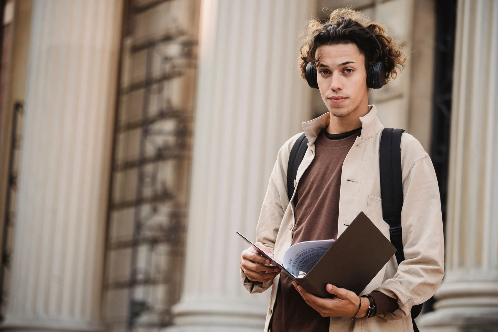 A young man with curly hair and headphones holding an open binder on a college campus