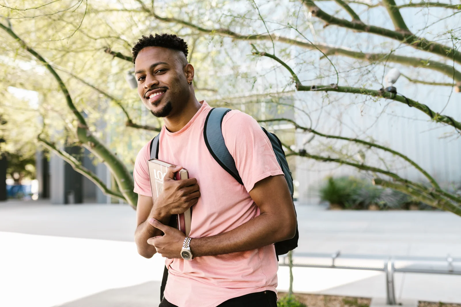 A smiling young man with a backpack holding a textbook on a college campus path