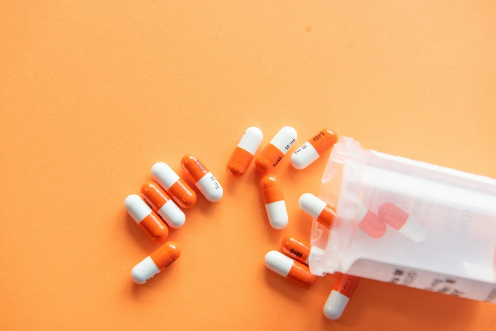 Orange and white capsules spilling from a pill bottle on an orange background