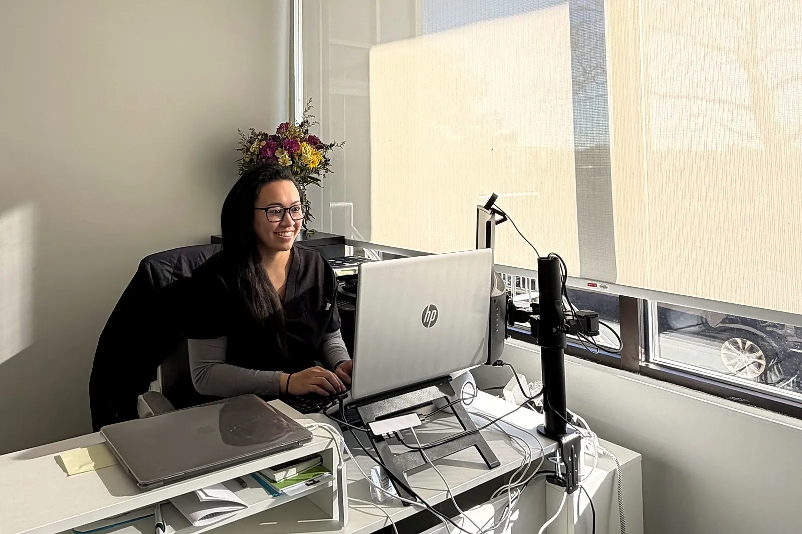 Kayla Yanga, NP, smiling at her desk in our Nutley office, with flowers on the windowsill behind her