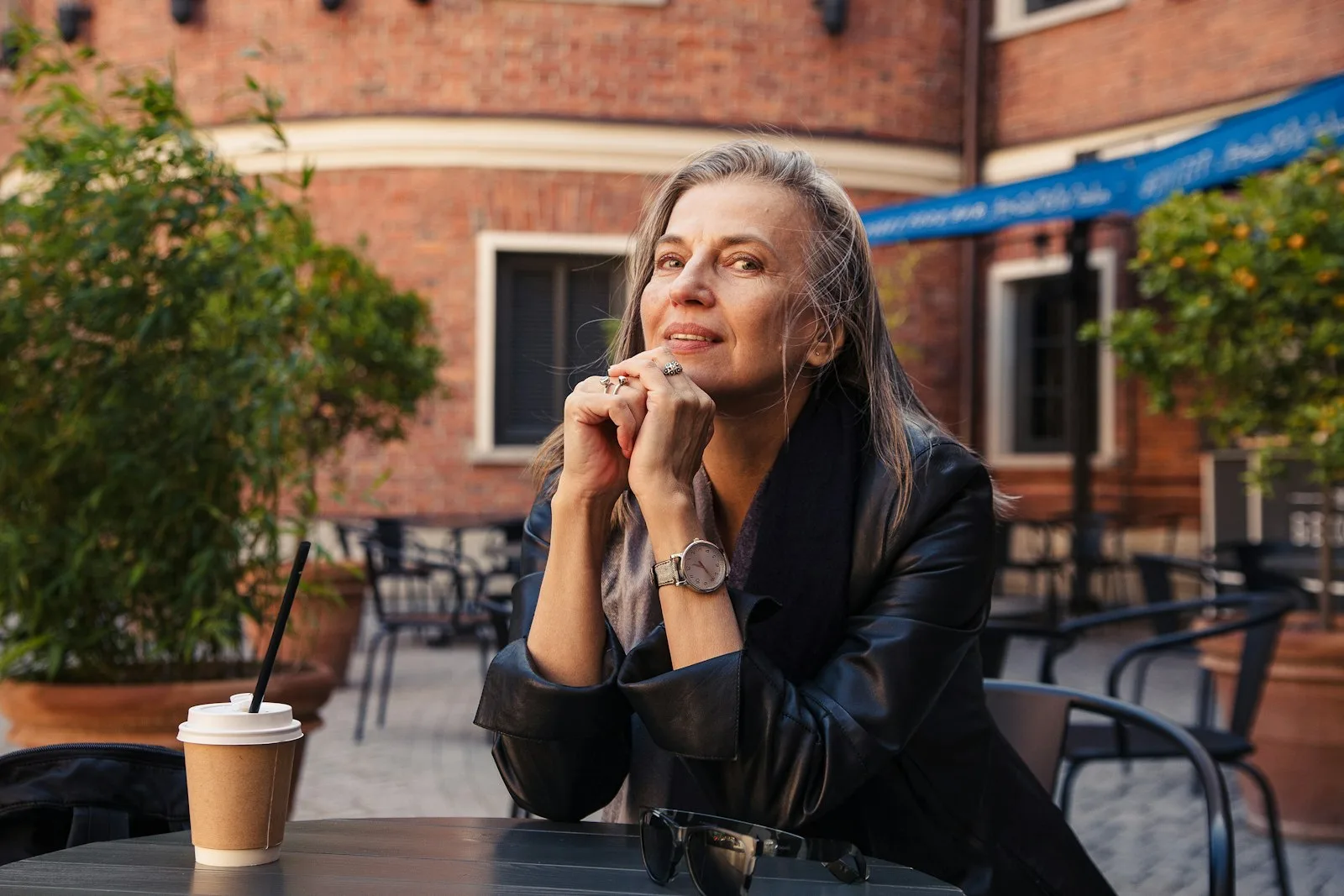 A woman with long gray hair sitting at an outdoor cafe, chin resting on her hands, looking thoughtful