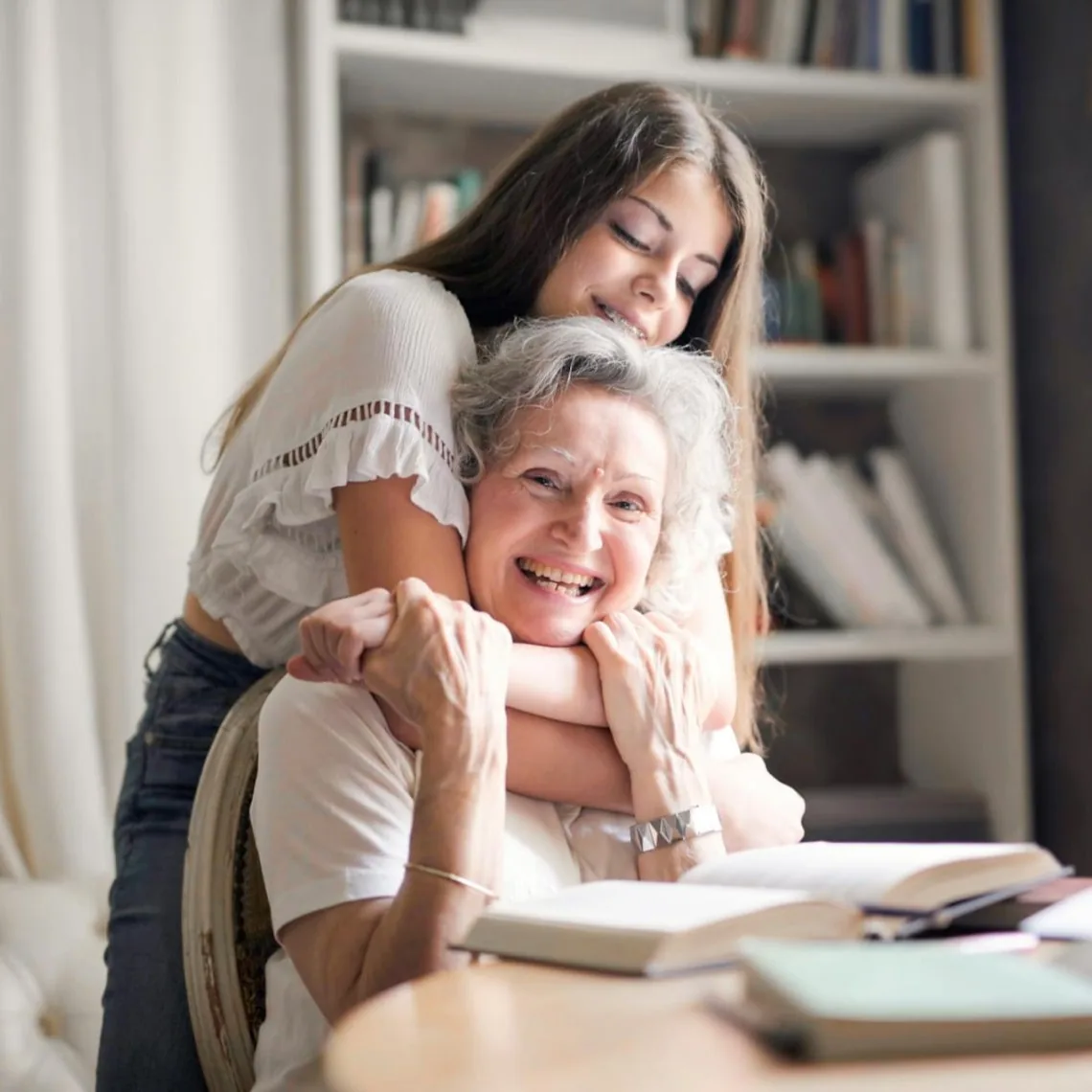 A young woman embracing her grandmother warmly at a bookshelf-lined desk