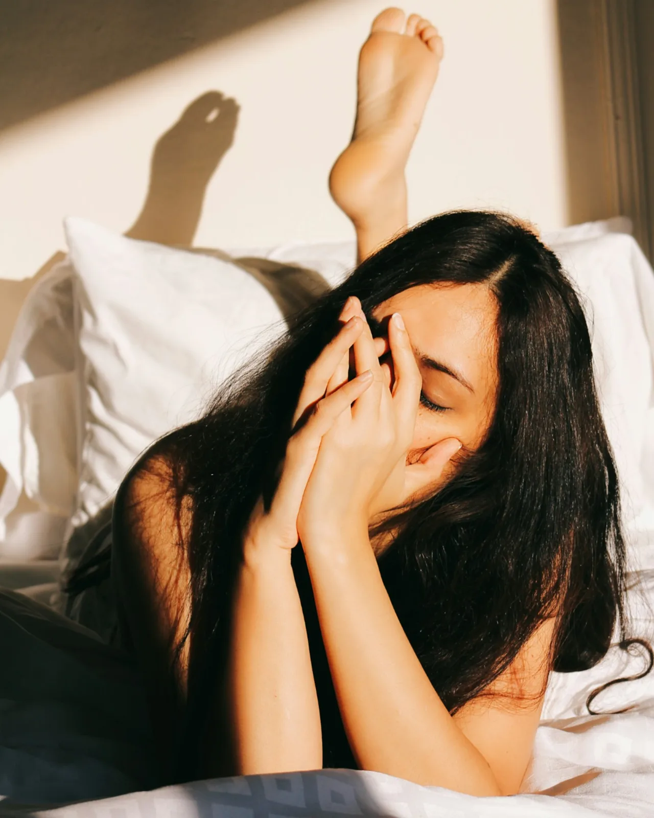 A woman lying in bed with her hands over her face in morning light