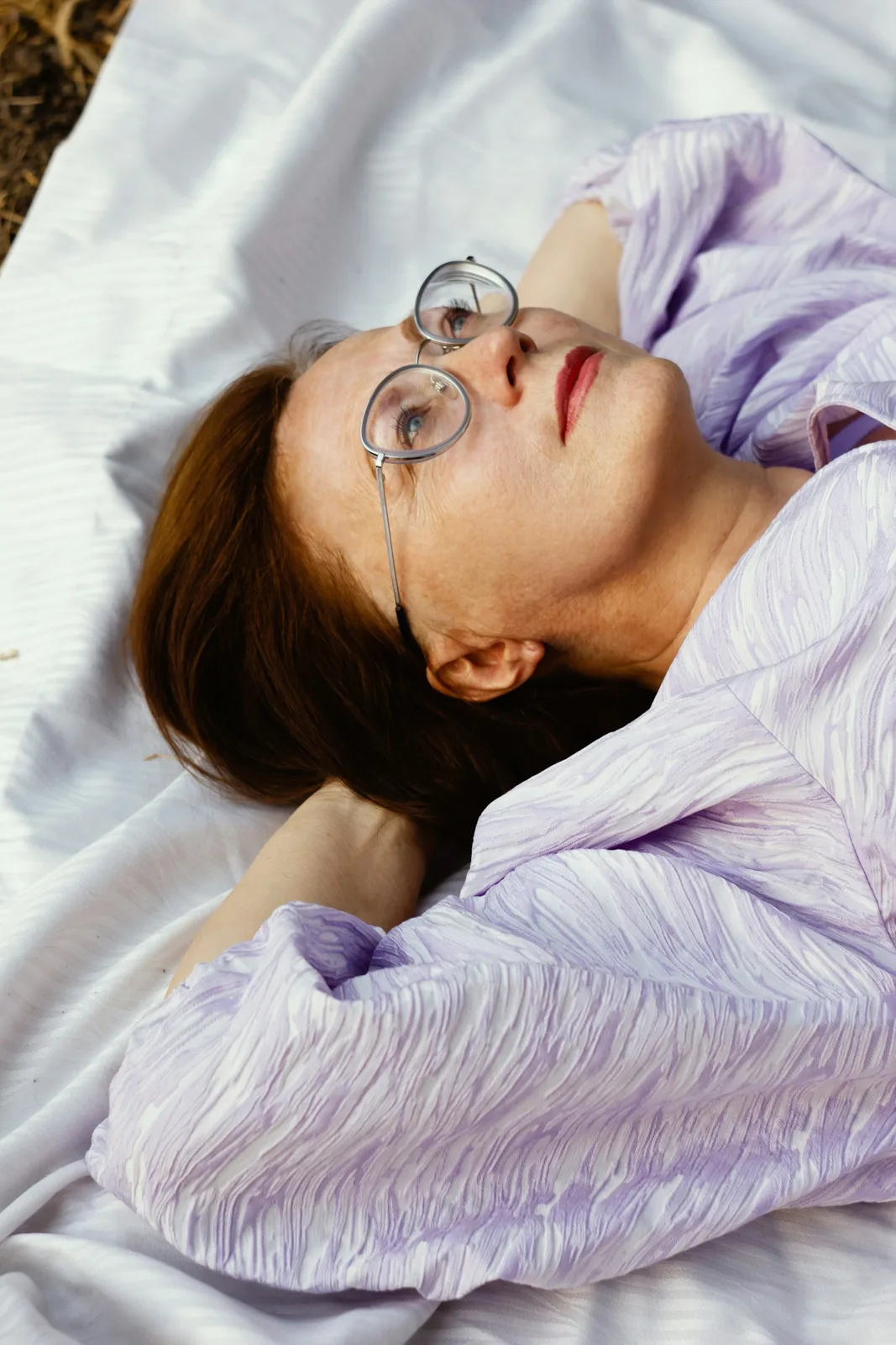 A woman with glasses lying on a soft blanket, gazing upward, lost in thought