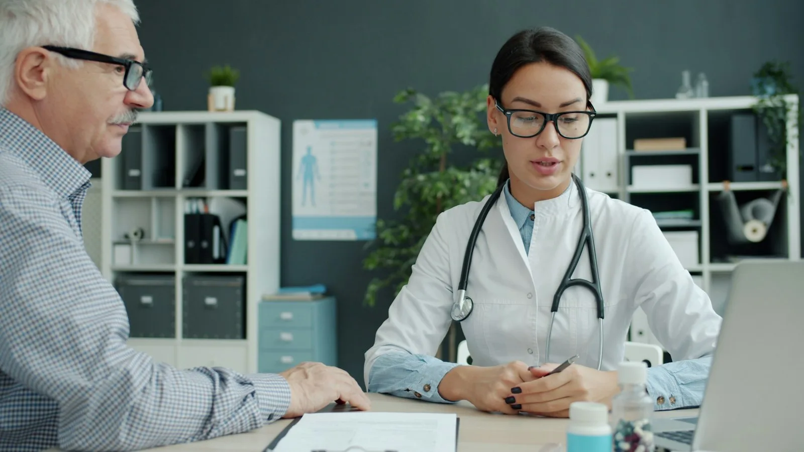 A clinician in a white coat with glasses reviewing notes at a desk with a patient